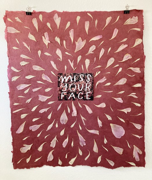 a postcard with white handwritten text "miss your face" on background of light pink mums centered on a large piece of burgundy lokta paper. light pink and cream-colored peony petals of various shapes and sizes are scattered all over the lokta paper.